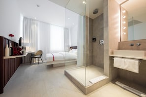 A modern hotel room with a glass-enclosed shower, a bed with white linens, a desk with a chair, and a vanity with a mirror. - Ruby Marie Hotel Vienna by IHG (Vienna)