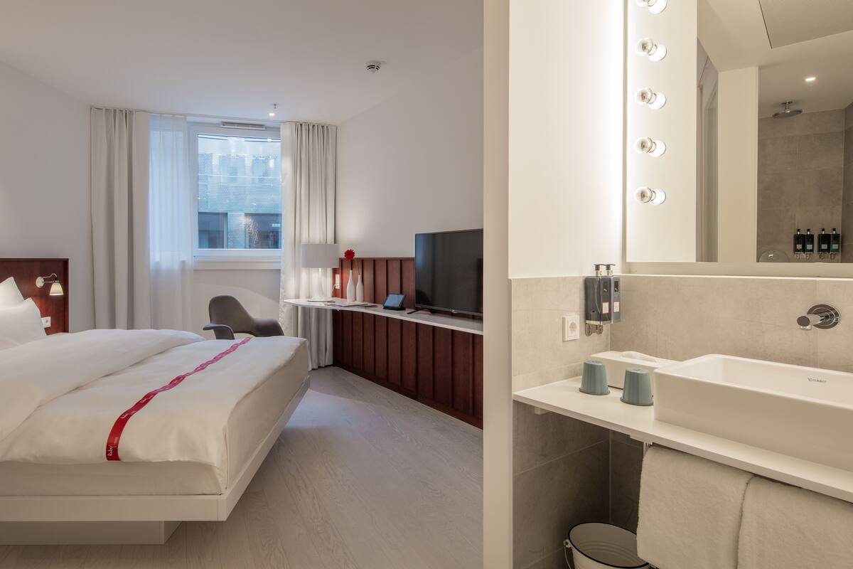A hotel room with a bed, a desk with a computer, a TV, a large mirror with lights, and a bathroom with a sink and shower.