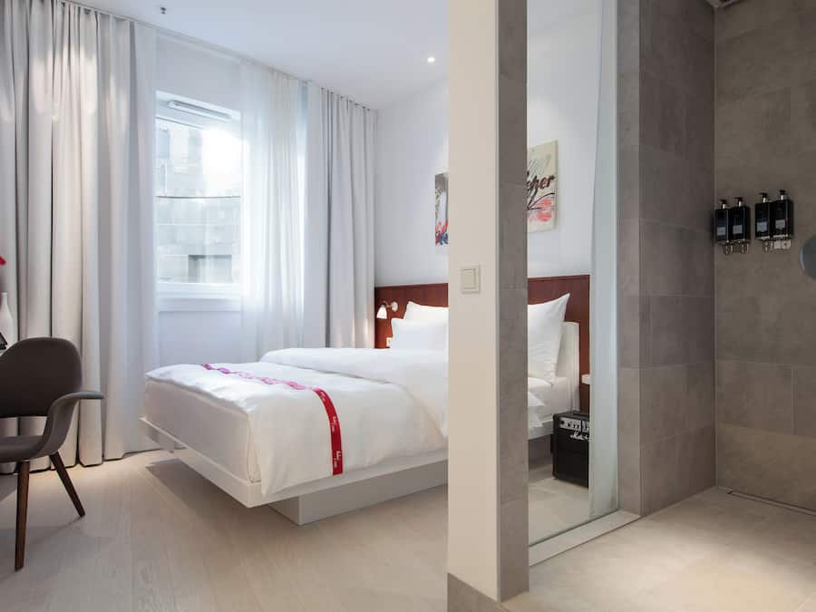 Habitación de hotel con cama, escritorio con computadora portátil, silla, zona de ducha con artículos de aseo y ventana con cortinas.