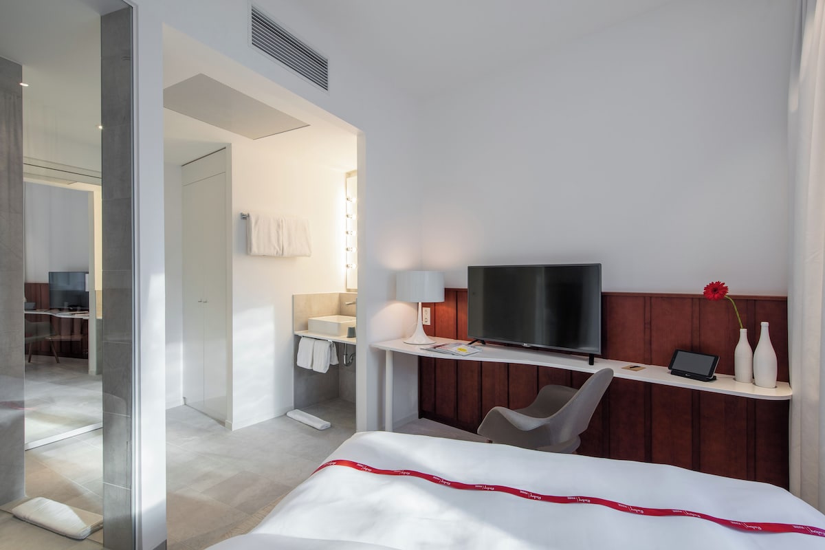 Una camera d'albergo con un letto, una scrivania con televisore, una sedia, un bagno con specchio e doccia.