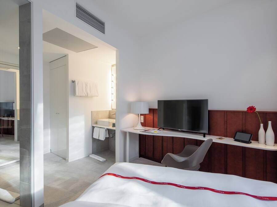 Habitación de hotel con cama, escritorio con televisor, una silla, baño con espejo y ducha.