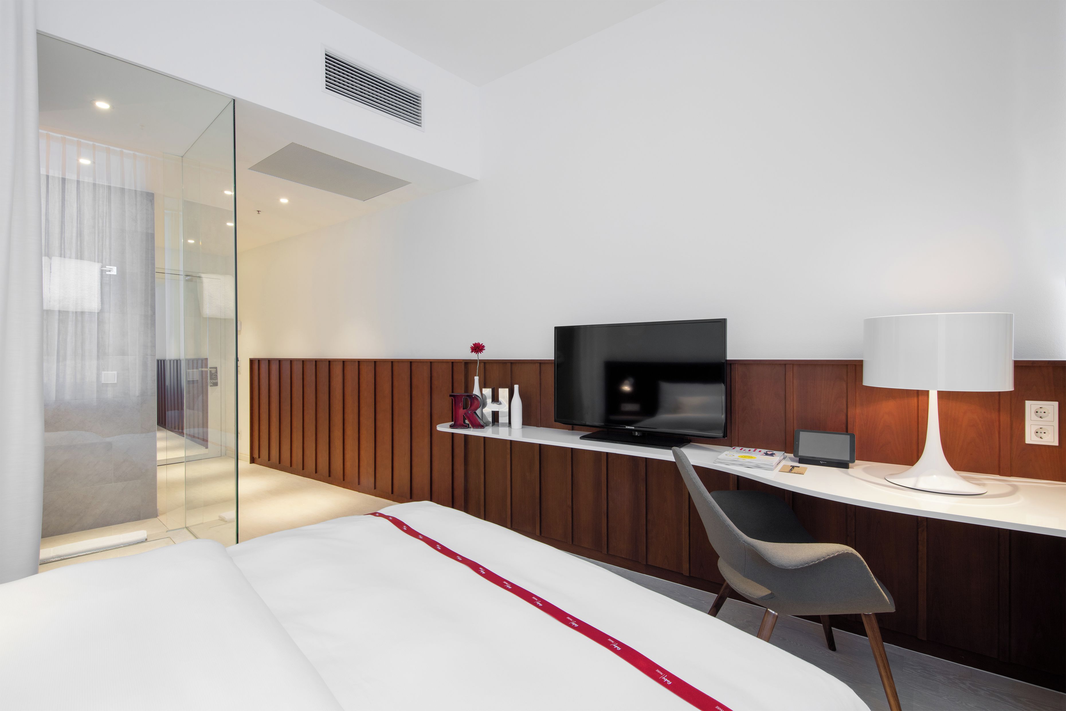 A modern hotel room with a bed, a desk with a television, a chair, and a bedside table with a lamp.