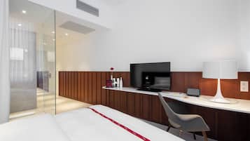 A modern hotel room with a bed, a desk with a television, a chair, and a bedside table with a lamp.