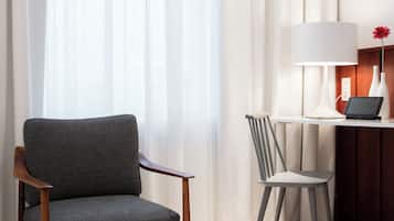 Habitación de hotel con una silla de madera, una silla más pequeña, un escritorio con una lámpara y un florero con una flor.