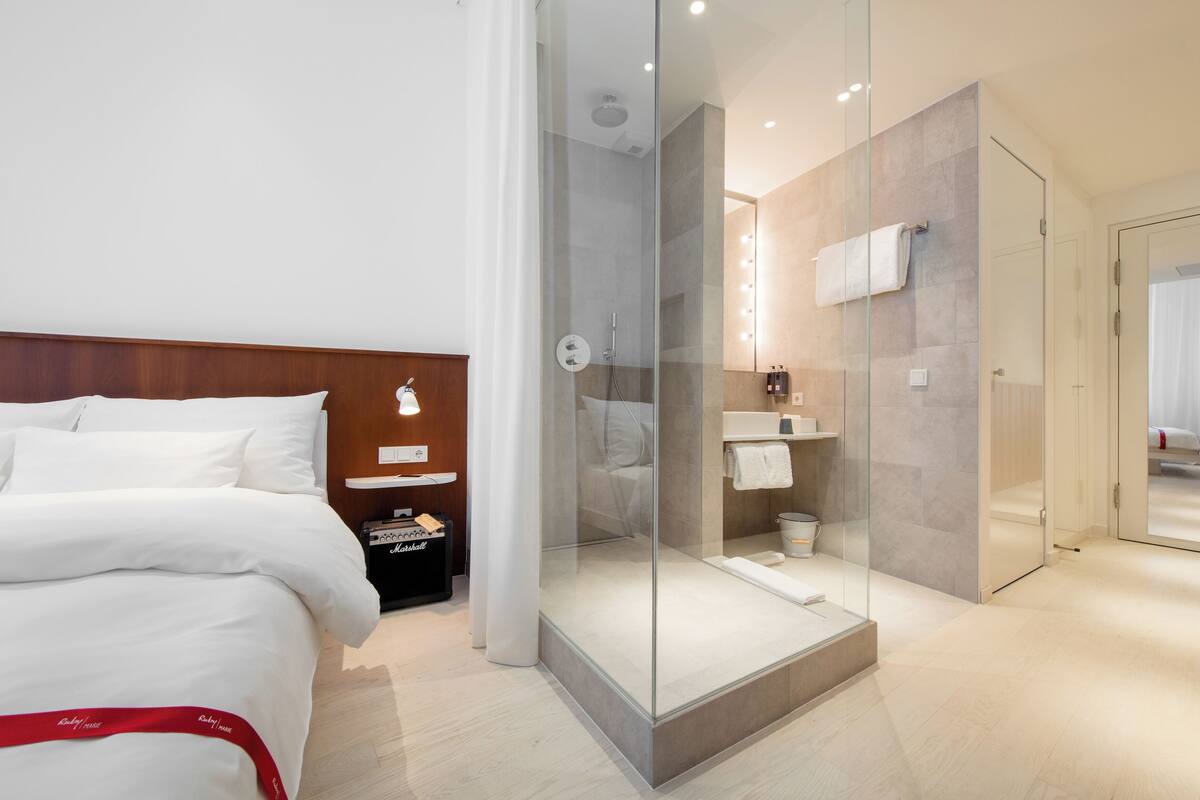 A modern hotel room with a bed, a glass-enclosed shower, a vanity with a mirror, and a red-striped bath mat.