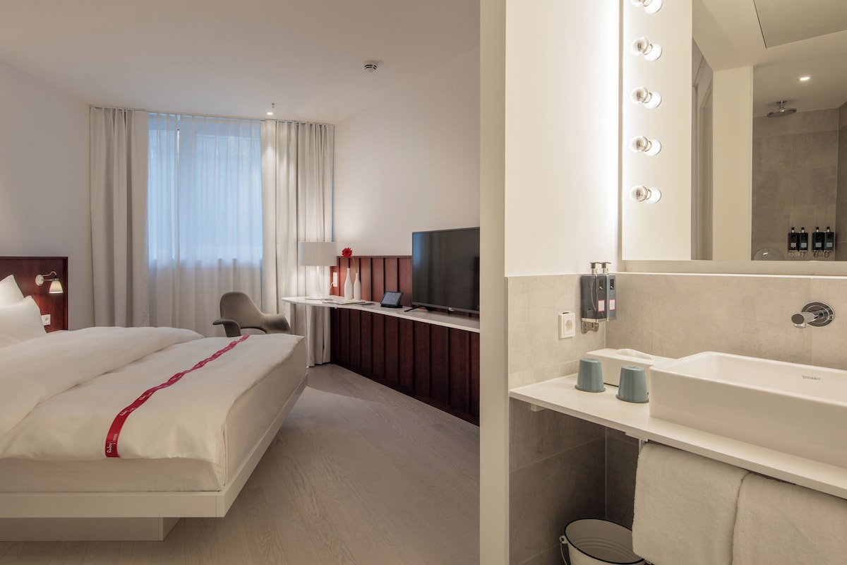 Una camera d'albergo moderna con un letto, una scrivania con televisore, uno specchio con luci e un bagno con lavandino e doccia.
