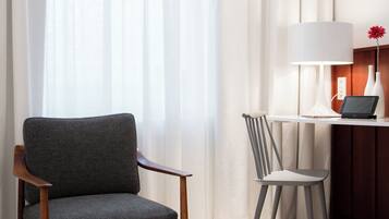 Quarto de hotel com uma cadeira de madeira, uma cadeira menor, uma escrivaninha com abajur e um vaso com uma flor.