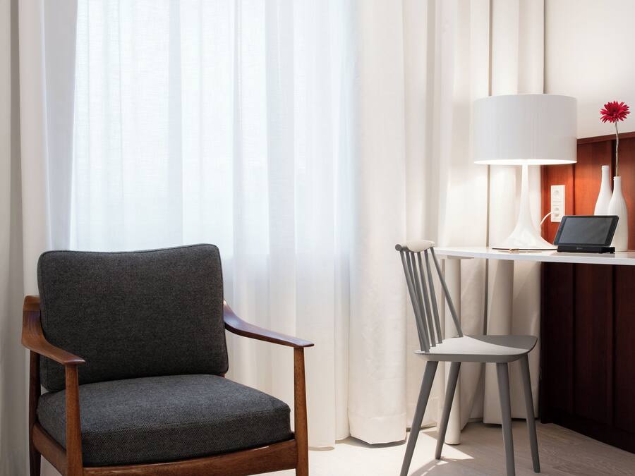 Quarto de hotel com uma cadeira de madeira, uma cadeira menor, uma escrivaninha com abajur e um vaso com uma flor.