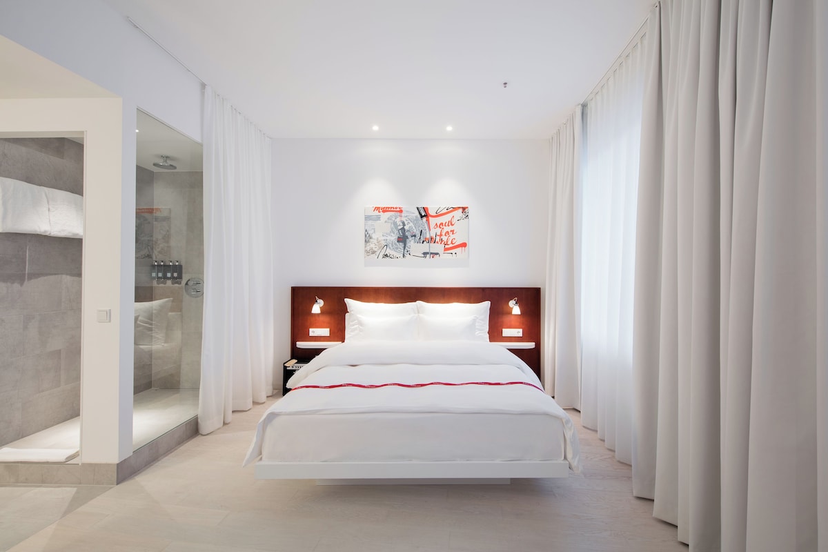 Una camera d'hotel moderna con un letto grande, uno spazio doccia in vetro e vista sulla città.