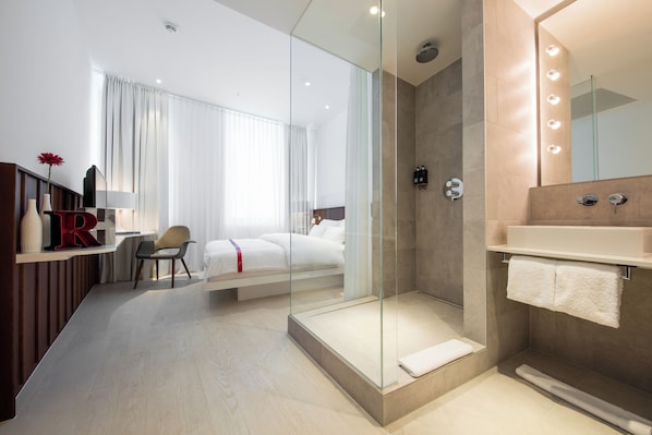 A modern hotel room with a glass-enclosed shower, a bed with white linens, a desk with a chair, and a vanity with a mirror. - Ruby Marie Hotel Vienna by IHG (Vienna)