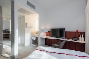 Una camera d'albergo con un letto, una scrivania con televisore, una sedia, un bagno con specchio e doccia.