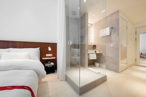 Una habitación de hotel moderna con cama, ducha con mampara de vidrio, tocador con espejo y una alfombra de baño a rayas rojas.