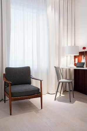 Una camera d'albergo con una sedia in legno, una poltroncina, una scrivania con una lampada e un vaso con un fiore.