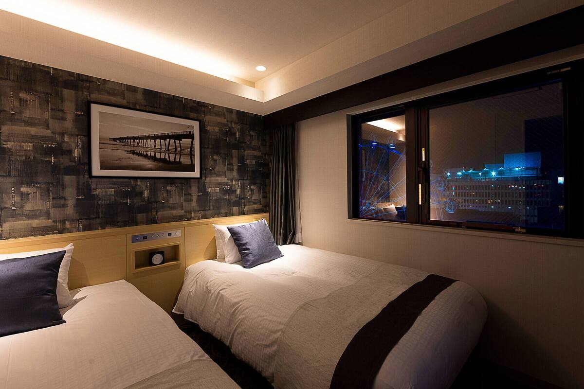 A hotel room with two beds, a nightstand, a clock, a framed picture on the wall, and a window with a city view at night.
