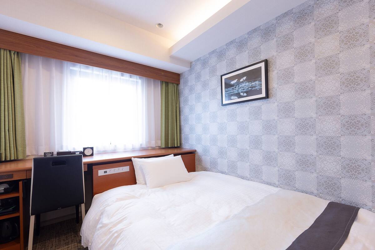 A hotel room with a bed, desk, and chair. There is a window with curtains, a wall with a patterned design, and a framed picture on the wall.
