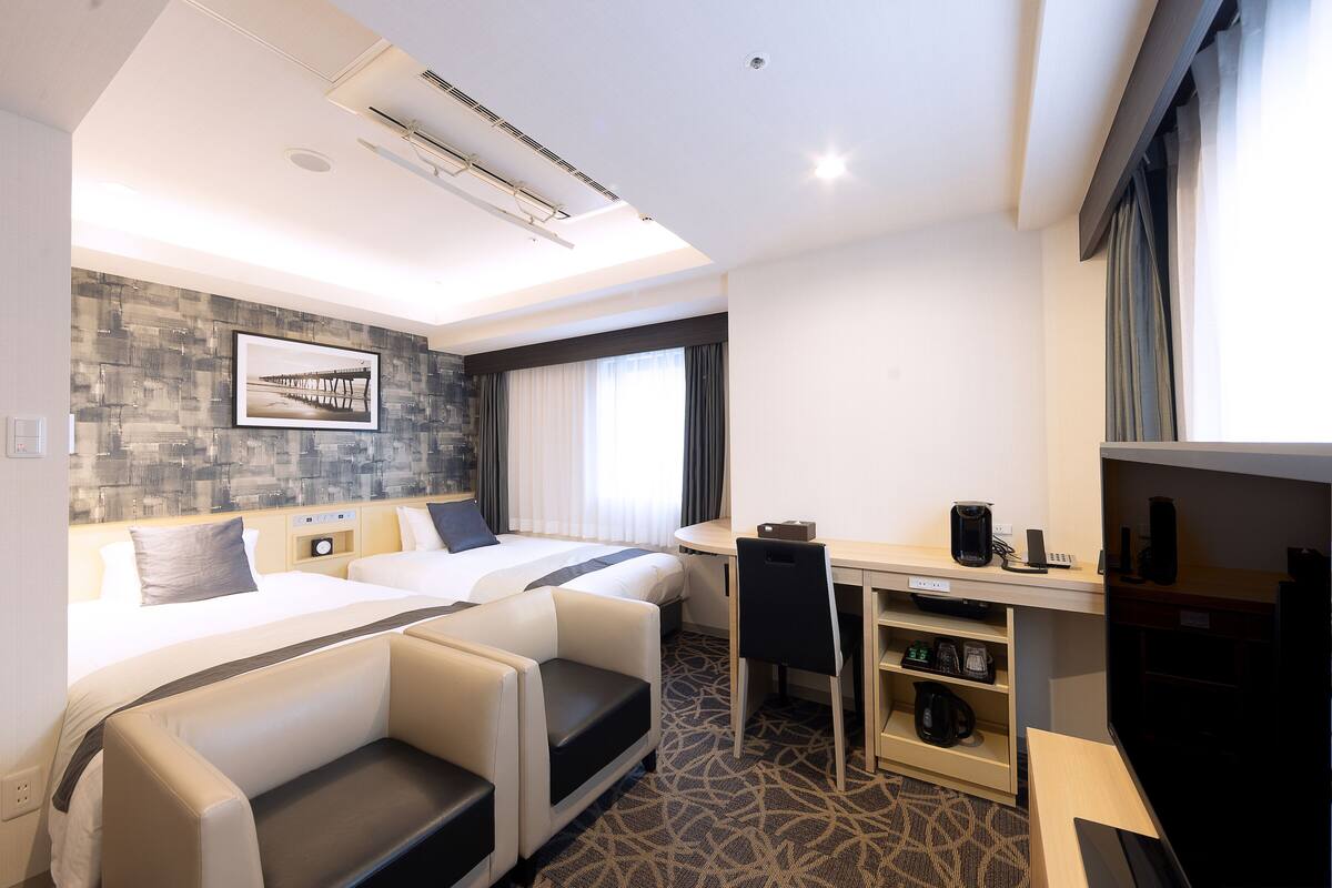 A hotel room with two beds, a desk, a chair, a TV, and a coffee machine.