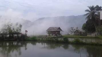 Una casa rustica su palafitte, accanto a un’acqua calma, circondata da vegetazione e con montagne nebbiose sullo sfondo.
