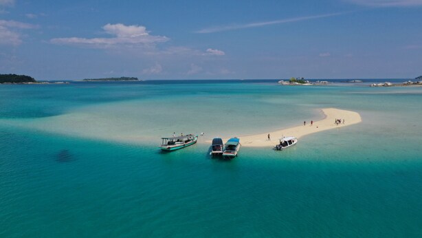 Une mer d’un bleu turquoise limpide, une plage de sable, des bateaux et une petite île au loin.