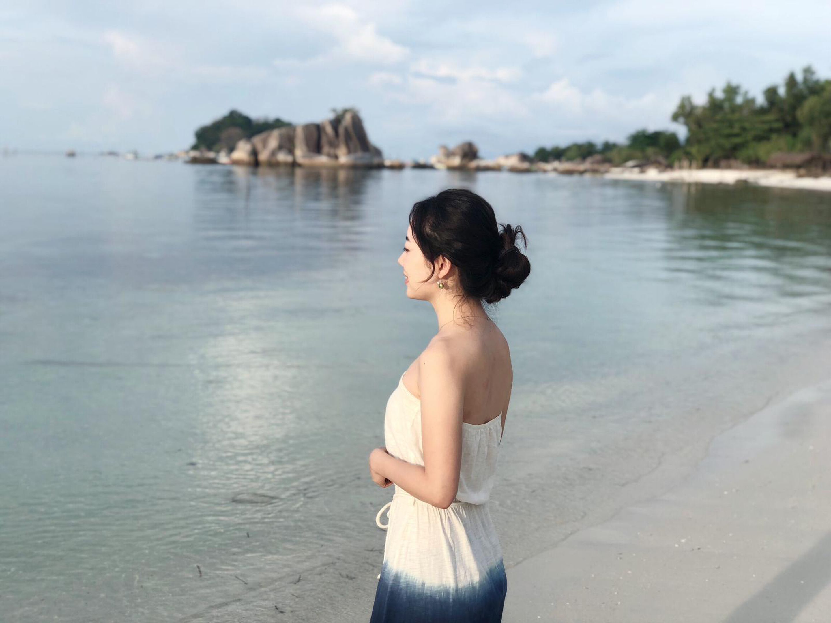 A woman standing on a beach with clear water and rocky formations in the background.