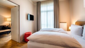 A hotel room with a bed, a TV mounted on the wall, a window with curtains, a lamp, and a red stool.