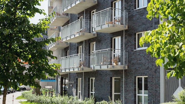 A multi-story residential building with balconies, a brick facade, and a small garden area.