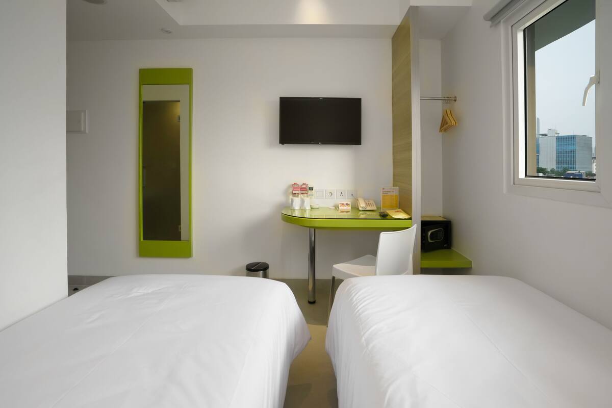 A hotel room with two beds, a TV mounted on the wall, a small table with toiletries, and a window with a city view.