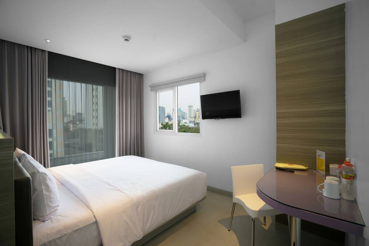A hotel room with a bed, a desk, a chair, a TV, and a window with a city view.