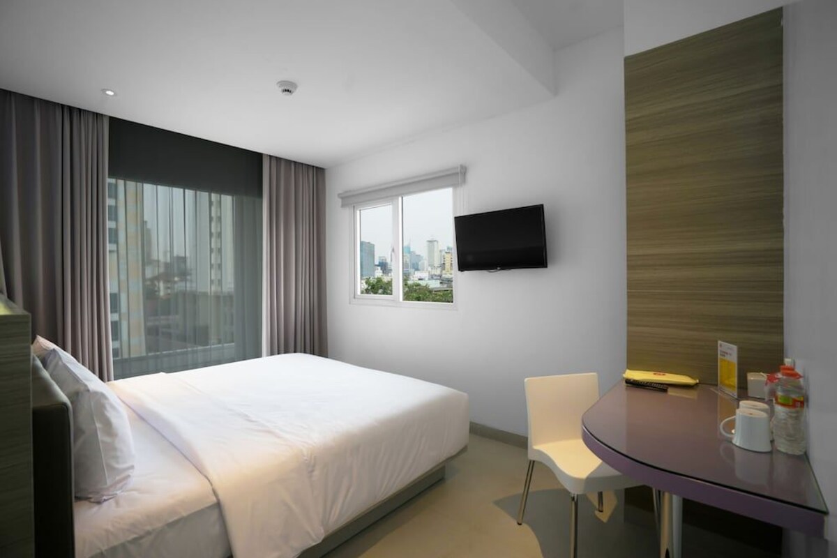 A modern hotel room with a large bed, a desk with a chair, a flat-screen TV mounted on the wall, and a window offering a city view.