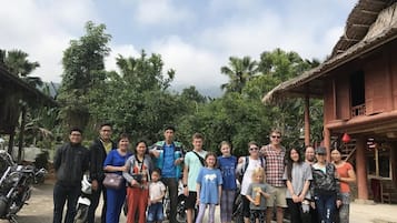 A group of people standing in front of a rustic building with a thatched roof, surrounded by greenery.