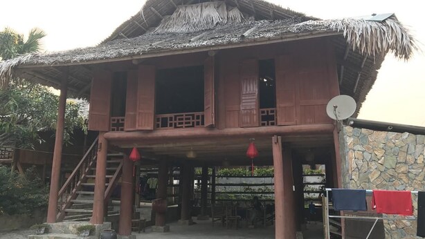 A traditional wooden house with a thatched roof, a staircase leading to an upper level, and a stone wall adjacent to the building. - Ban Buoc Homestay - Hostel (Muong Bi)