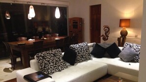 A modern living room with a white sofa, patterned cushions, a dining table, and pendant lighting. - Villa Pantai Lovina (Kubutambahan)