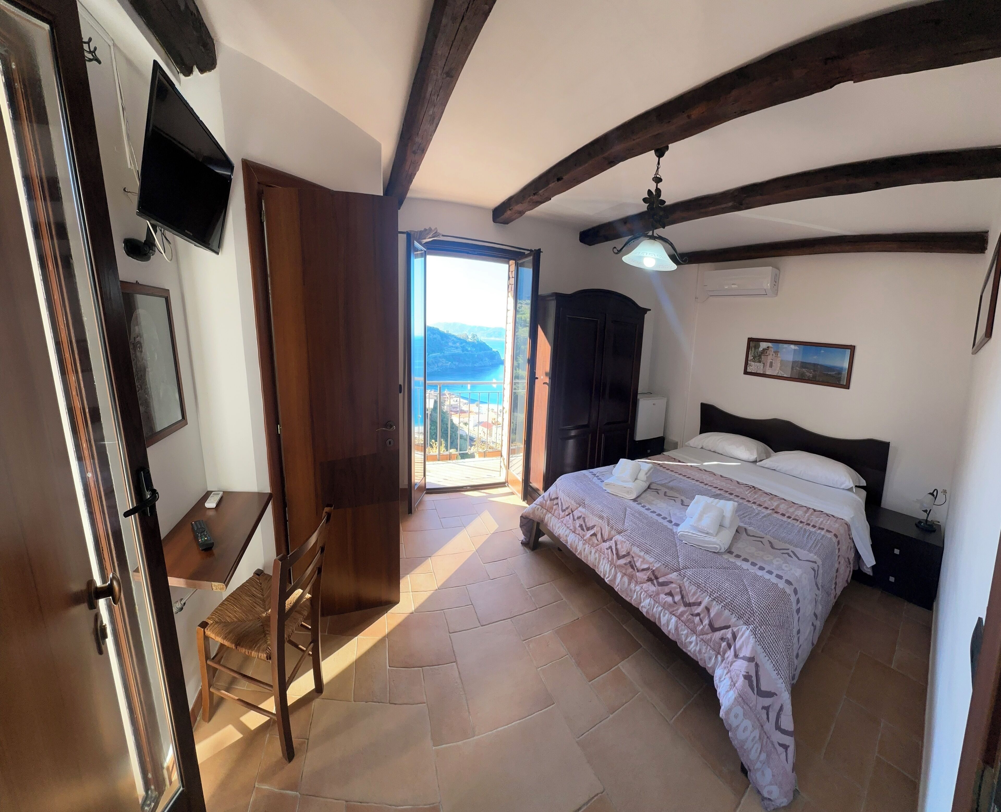 A bedroom with a bed, a desk, a chair, a wardrobe, and a balcony with a view.