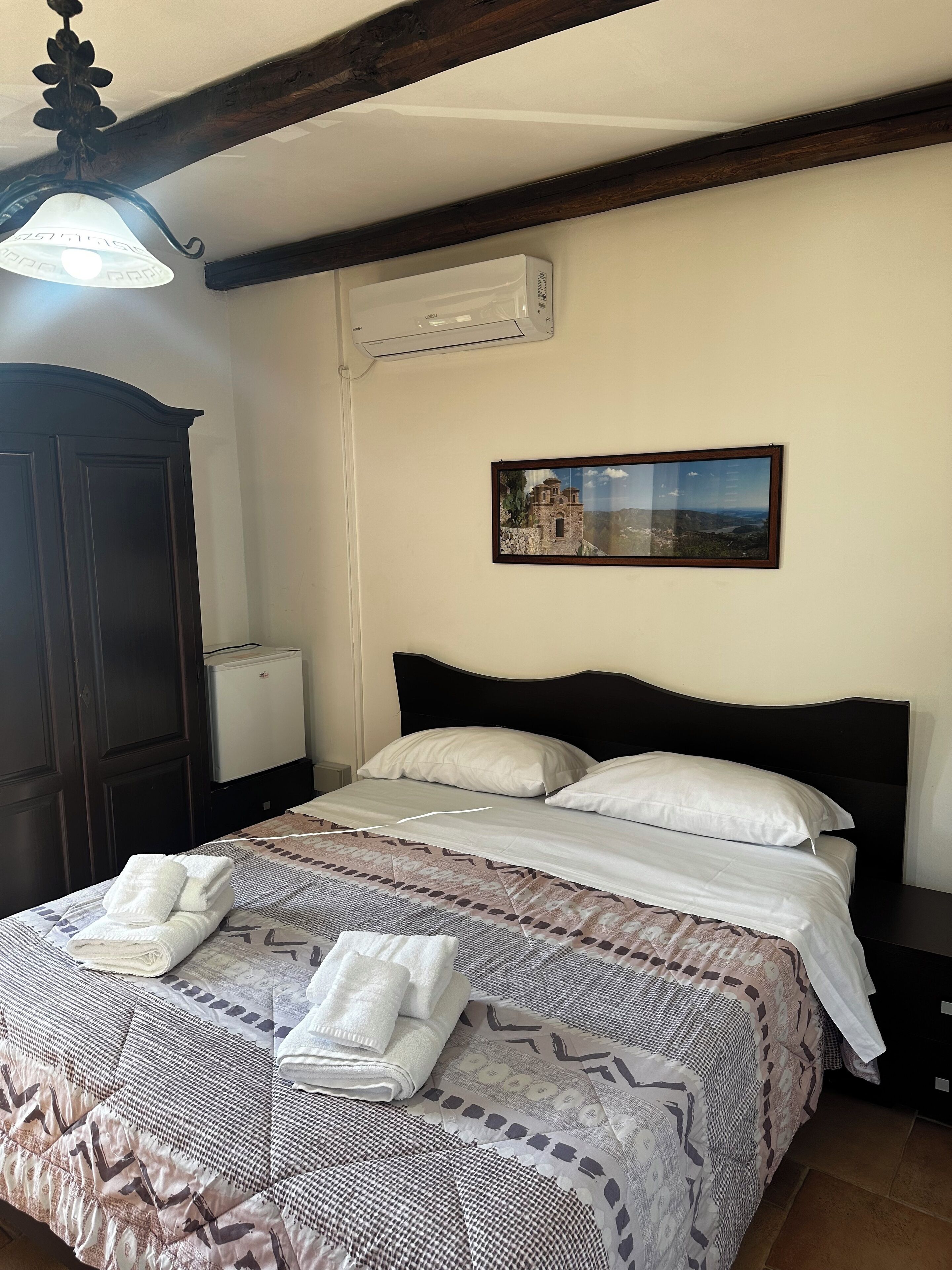 Una cama bien hecha con una colcha estampada y dos almohadas, un cabecero de madera oscura, un aire acondicionado de pared y un cuadro enmarcado con un paisaje.