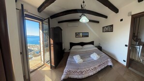 A bedroom with a bed, a nightstand, a hanging light fixture, a window with a view of the sea, and a door leading to a balcony. - B&B La Bastia  (Scilla)