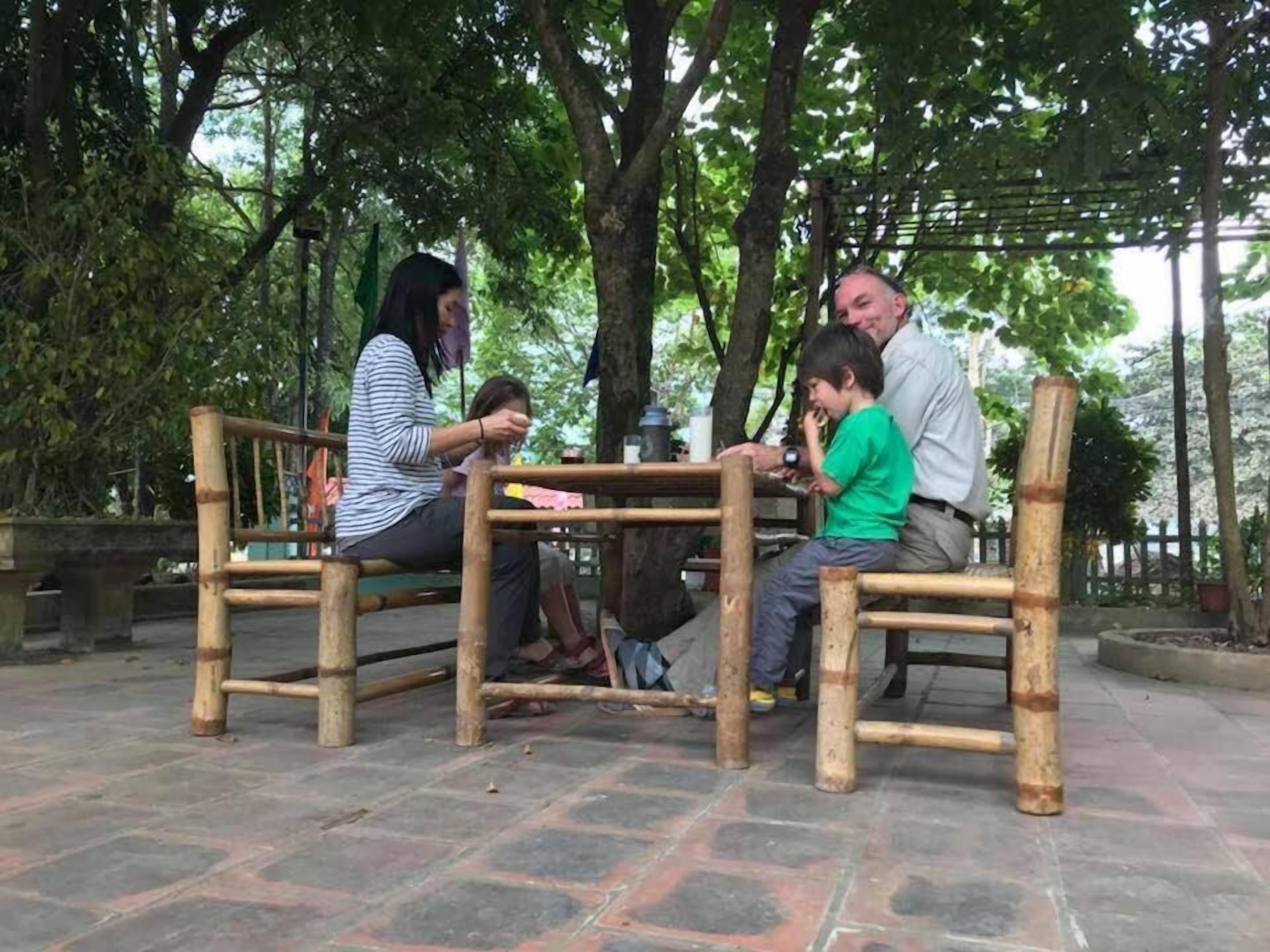 A group of people seated at a bamboo table in an outdoor setting with trees in the background.