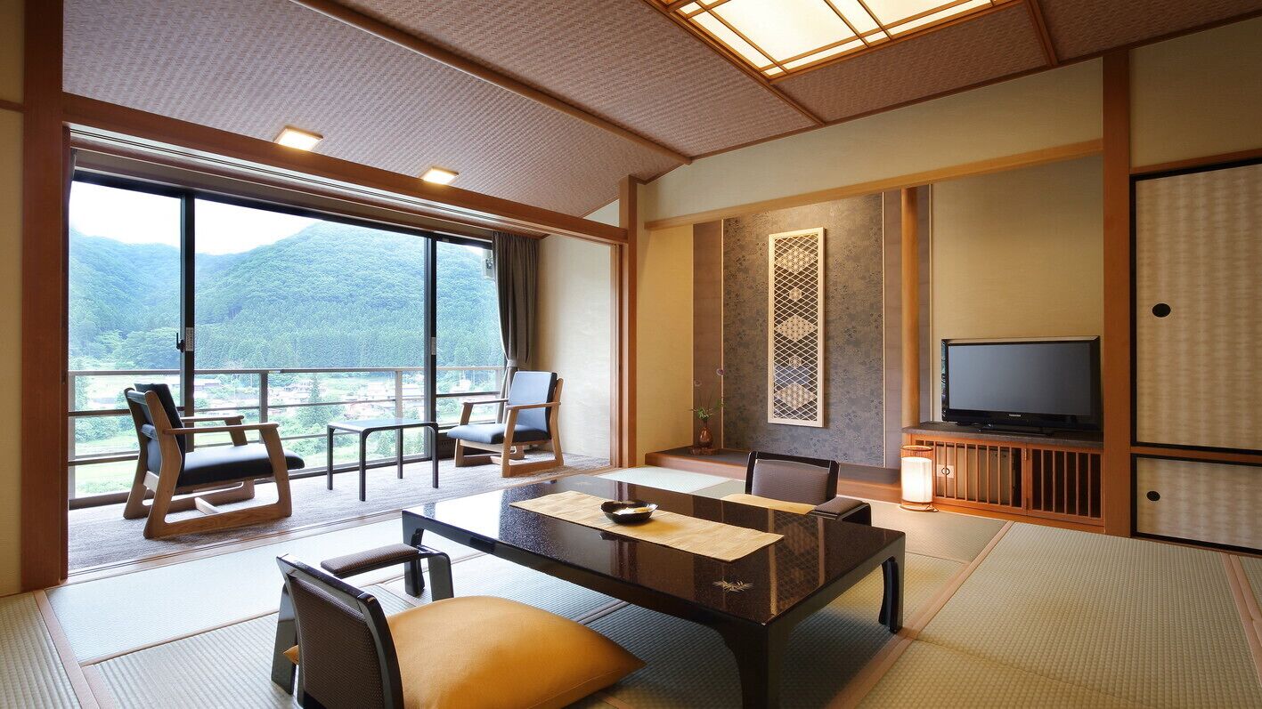 A traditional Japanese room with tatami flooring, a low table, chairs, a television, and a view of a mountainous landscape.