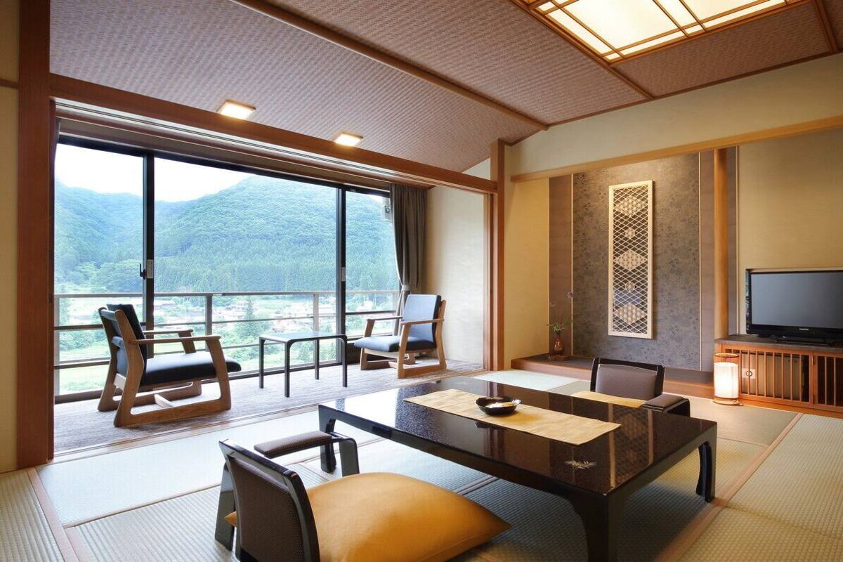 Une pièce de style japonais traditionnel, avec un sol recouvert de tatamis, une table basse, des chaises, un téléviseur et une vue sur un paysage montagneux.