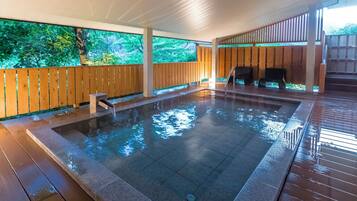 A rectangular indoor swimming pool with a wooden deck surrounding it, a wooden fence, and a view of greenery outside.