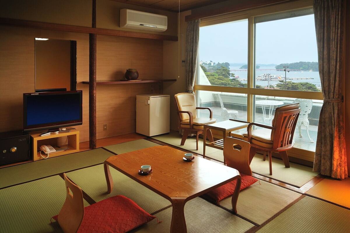 A room with tatami mats, a television, a refrigerator, a table with chairs, and a view of a body of water through large windows.