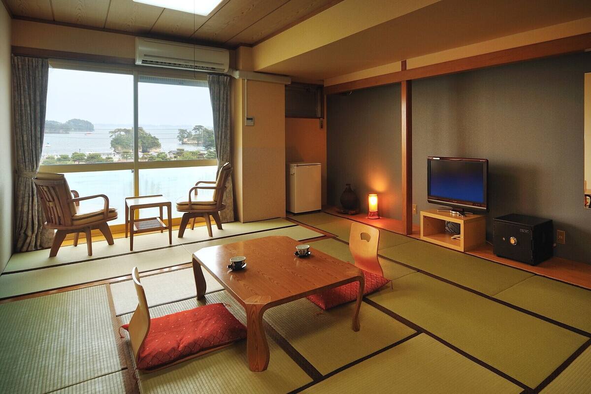 A traditional Japanese-style room with tatami flooring, a television, a small table, and a view of the sea.