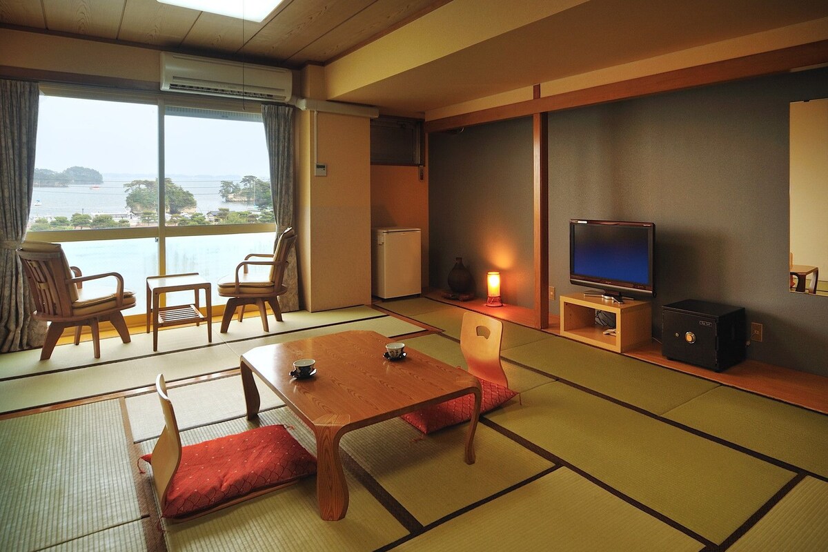 A traditional Japanese-style room with tatami flooring, a television, a small table, and a view of the sea.