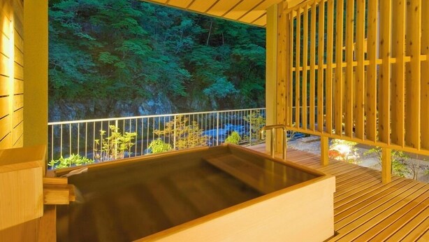 A wooden deck with a hot tub, a wooden railing, and a backdrop of a forest scene. - Shofurou Matsuya (Nasushiobara)