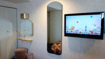 A bathroom with a mirror, a television mounted on the wall, a small shelf, and a patterned ottoman.
