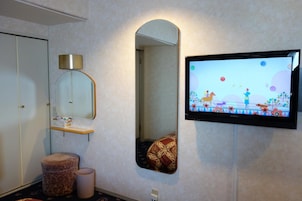 Un baño con espejo, televisor montado en la pared, estante pequeño y otomana estampada.