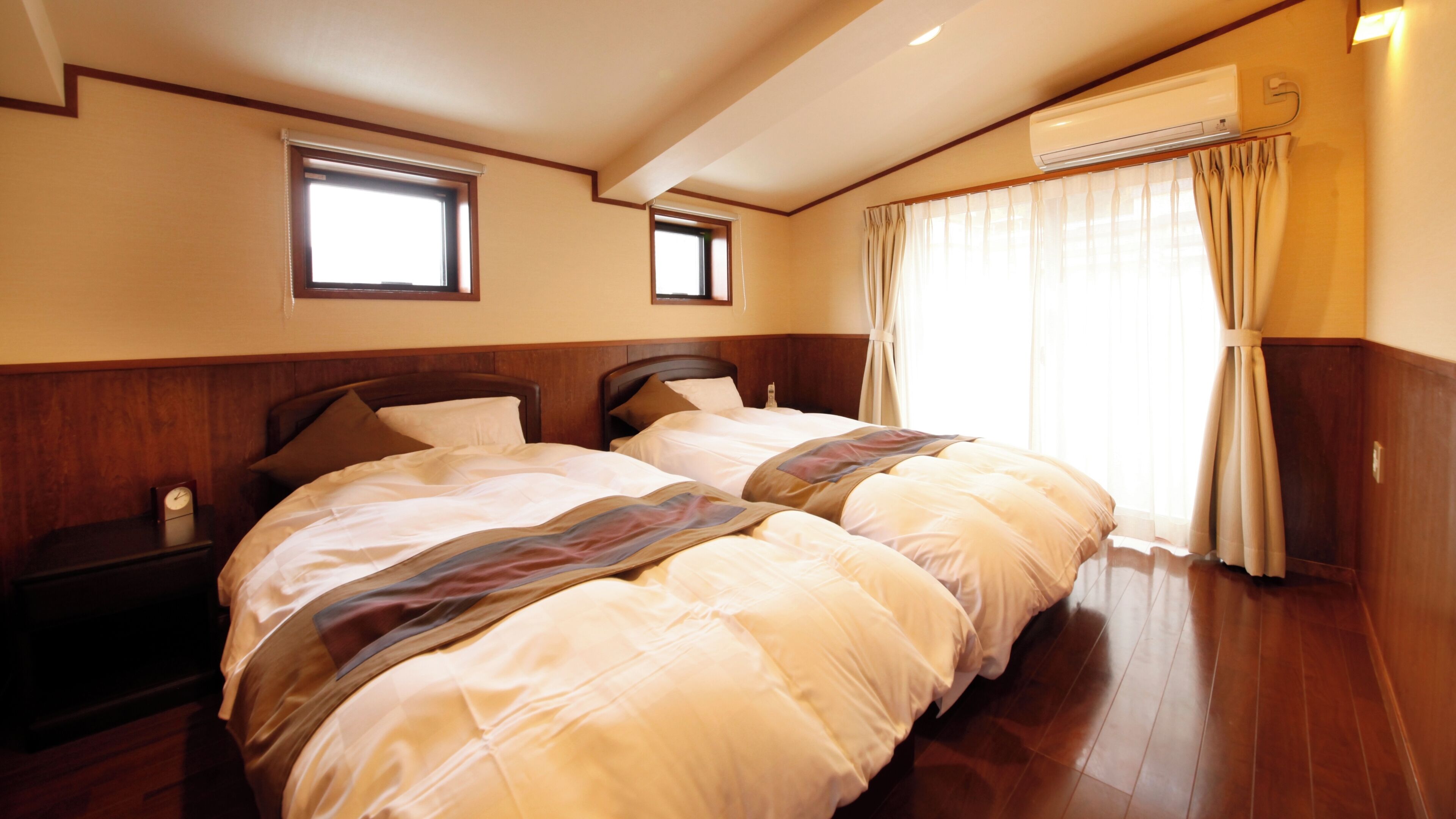 A hotel room with two beds, wooden paneling, an air conditioning unit, a window with curtains, and a clock on a nightstand.