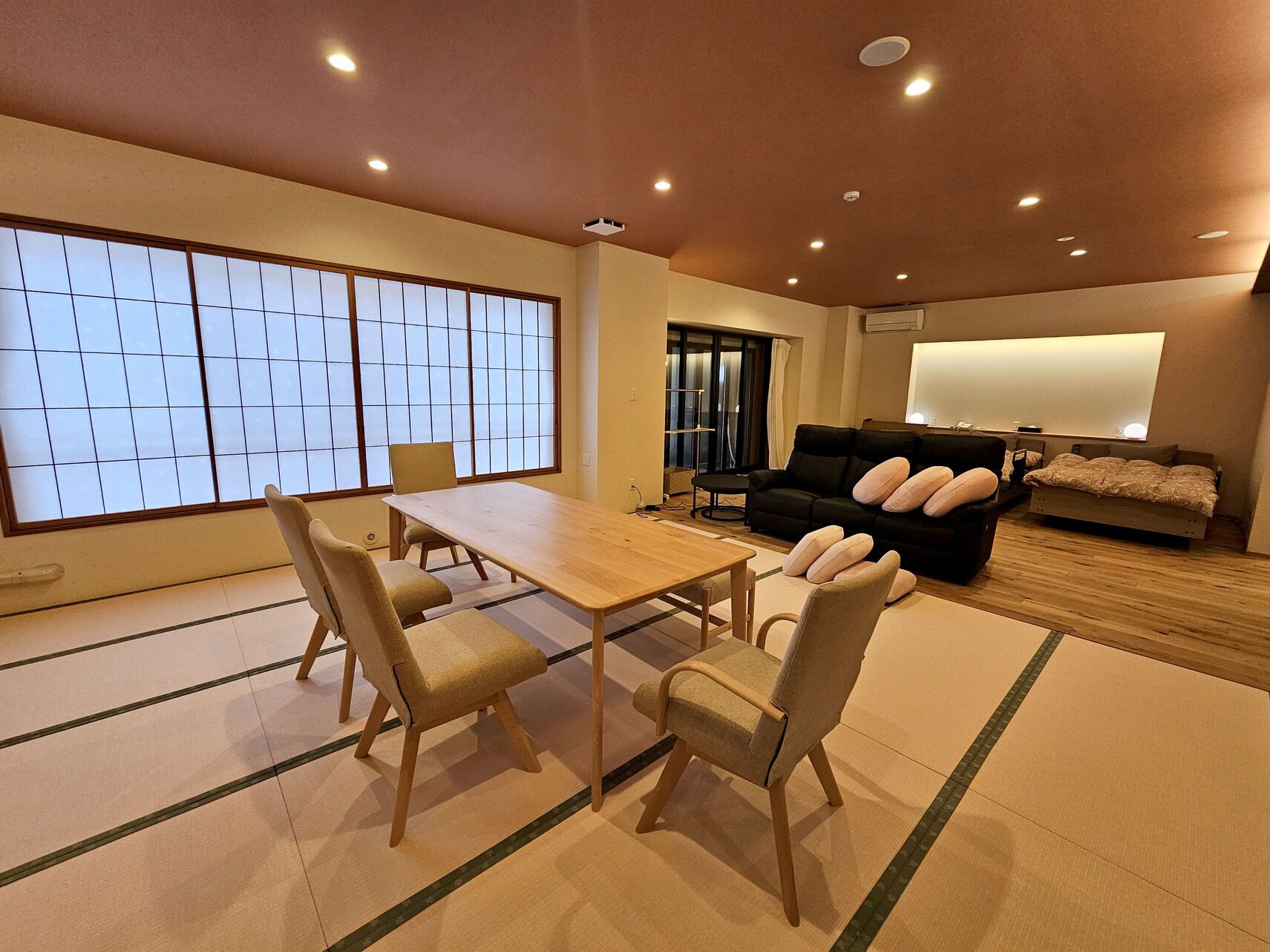 A room with tatami mats, a wooden table, chairs, a black sofa, and a bed with pillows.
