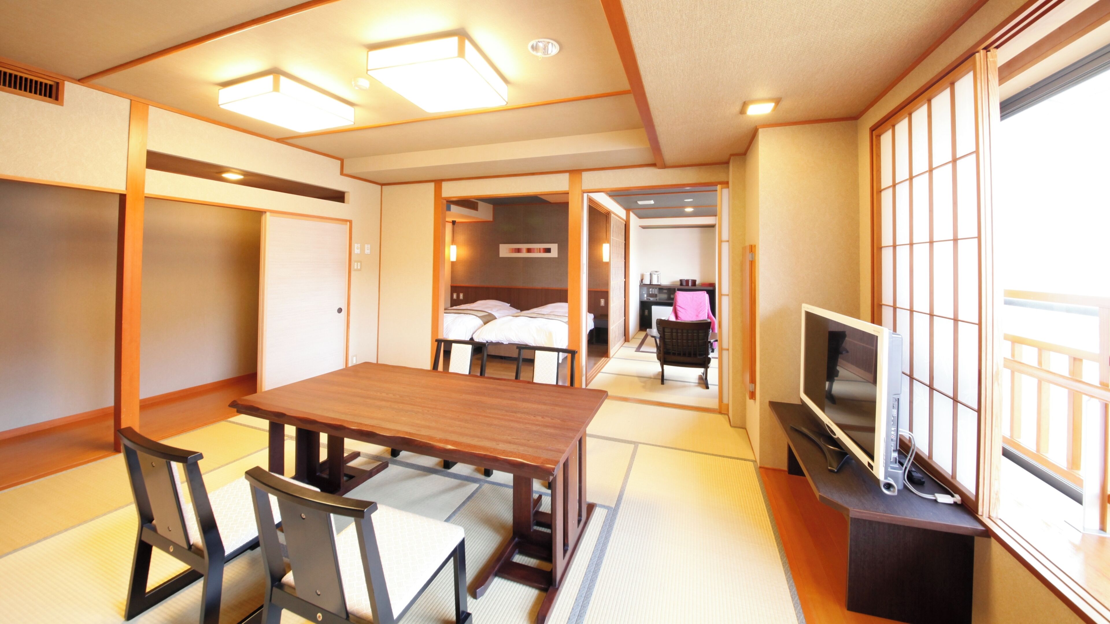 A traditional Japanese room with tatami flooring, a wooden table, chairs, a television, and a sliding door leading to a bedroom with a bed.