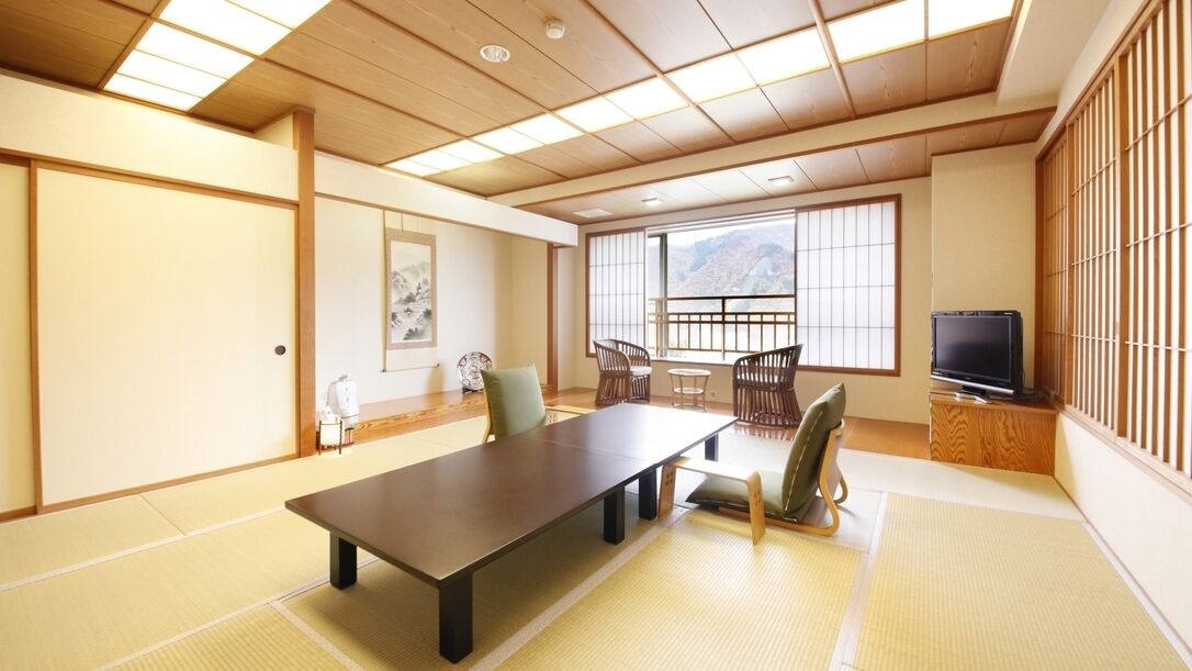 A traditional Japanese room with tatami flooring, a low table, chairs, a television, and a view of mountains through large windows.