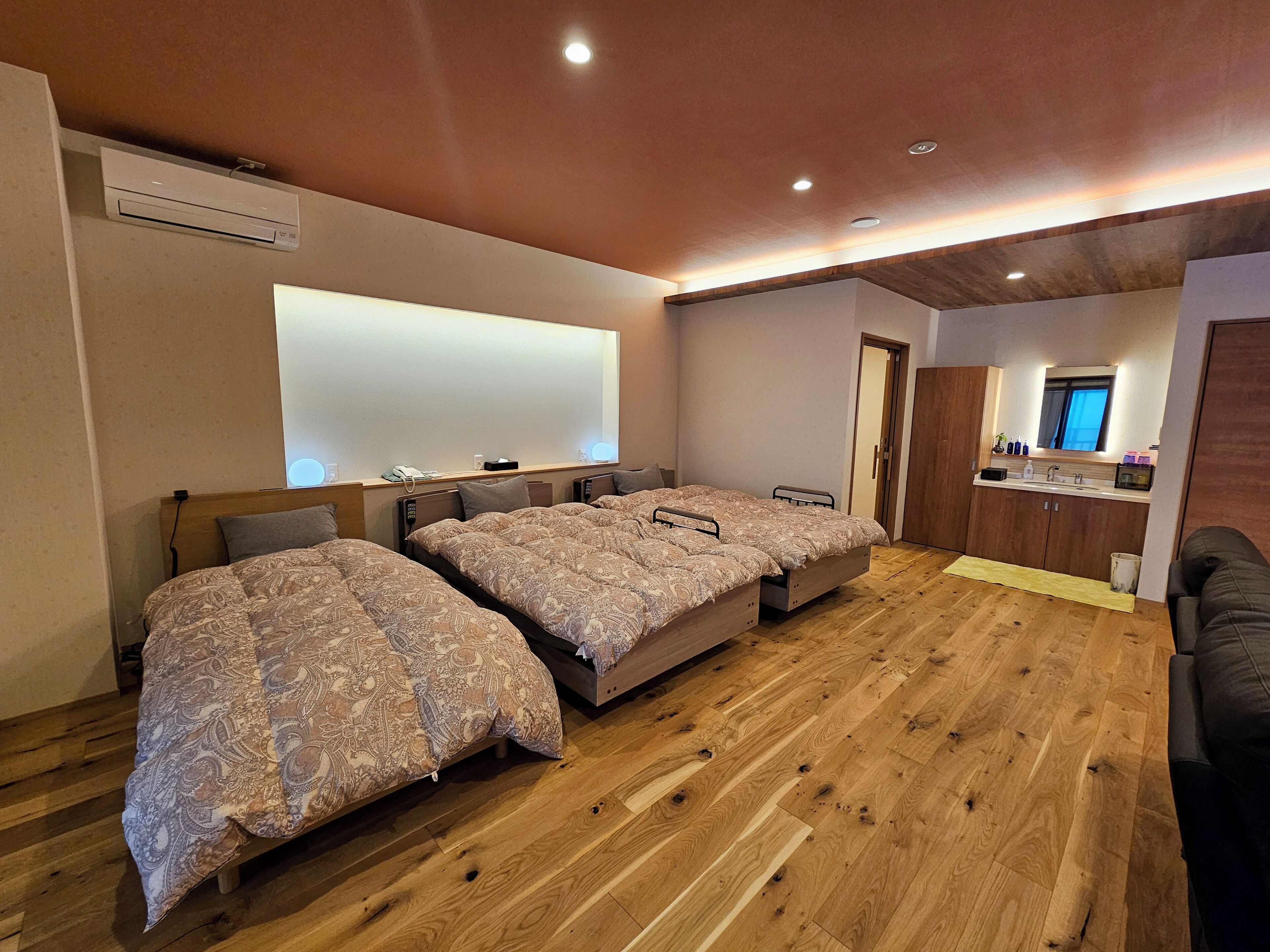 A hotel room with three beds, a wooden floor, a large mirror, and a kitchenette.