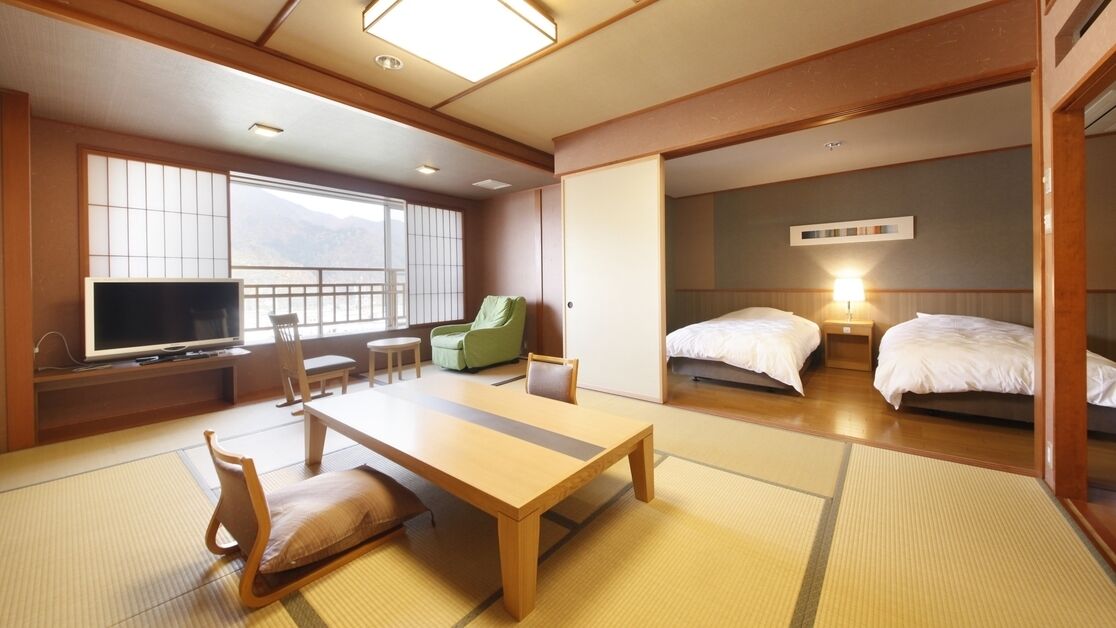 A traditional Japanese-style hotel room with tatami flooring, a low table, a green chair, two beds, and a television.